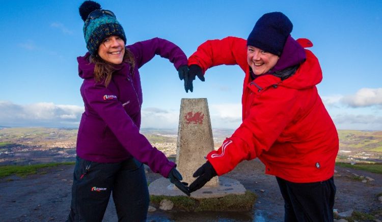 Two ramblers making a heart with their arms around a Welsh waymarker at a high elevation