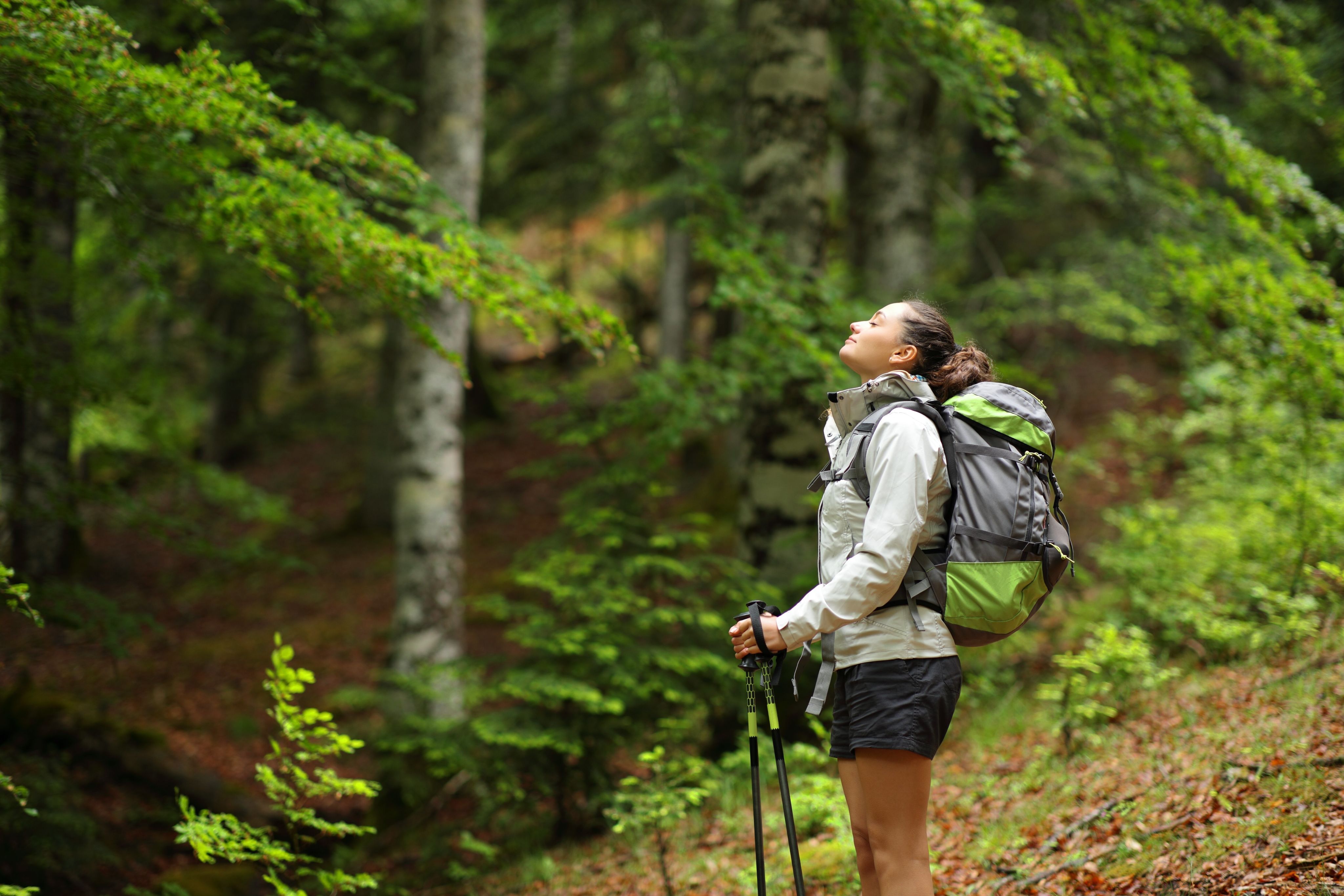 Woman with poles and rucksack standing in woods with her eyes closed and smiling